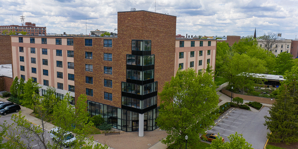 A brick building with glass windows. A parking lot is to the left and green trees are in the front. 